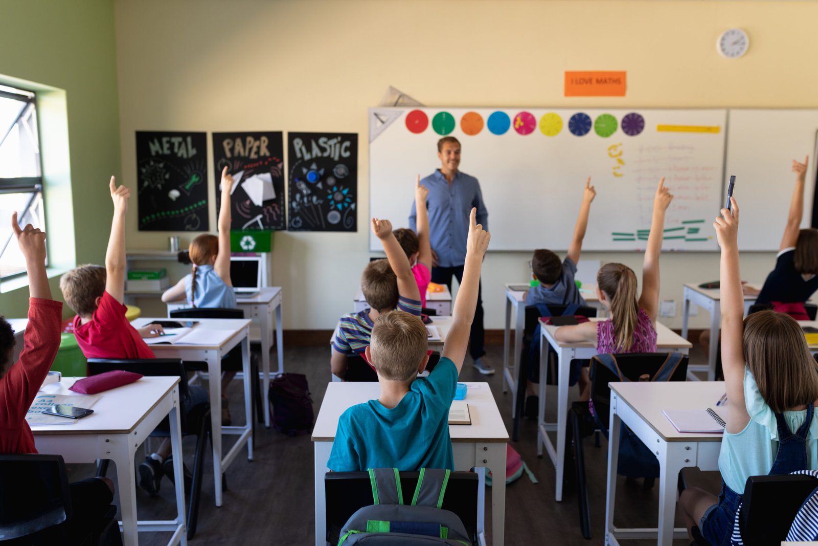 Front view of a Caucasian male school teacher standing in front of a diverse group of school children sitting at desks and raising their hands to answer a question during a lesson in an elementary school classroom